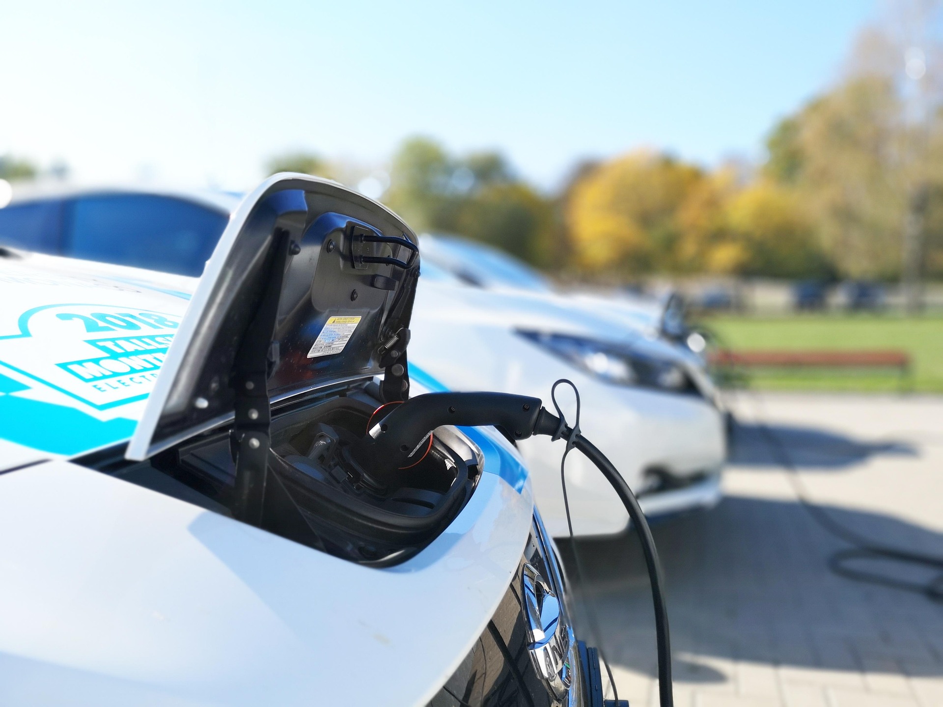 A white electric car with its charge-port door open and a J1772 connector plugged in, with a second EV charging behind it.