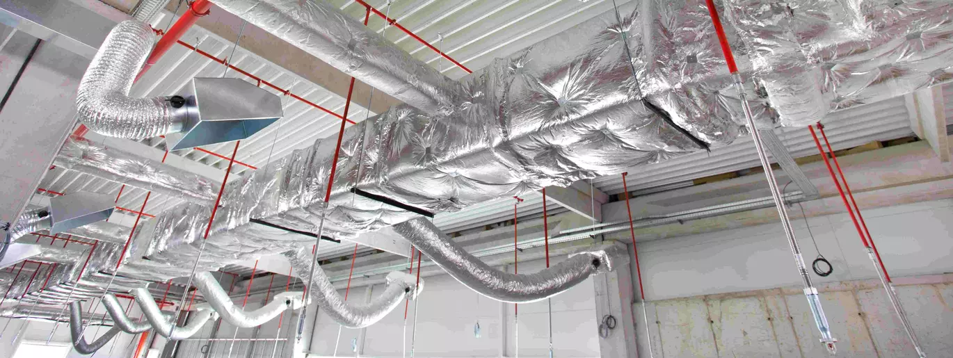Insulated flexible duct branching off a rectangular sheet-metal trunk in an exposed ceiling, suspended on hanger straps alongside red sprinkler piping.